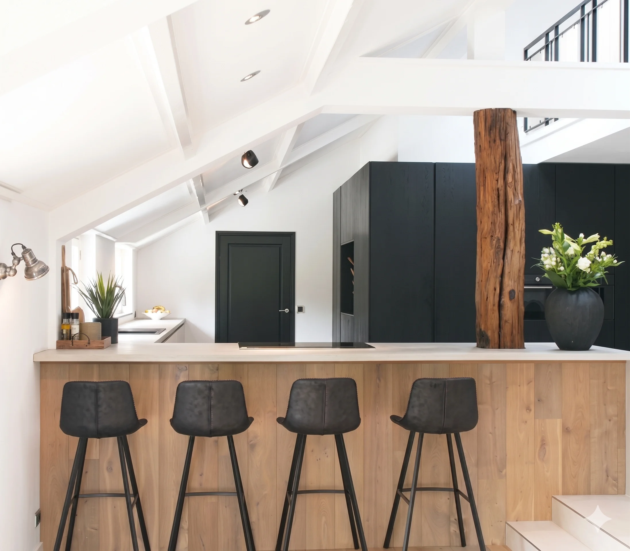 Moderne robuuste keuken met een hellend plafond, strakke zwarte kastenwand en een sfeervolle houten bar met kookplaat en barkrukken.